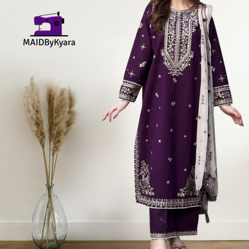May include: A deep purple embroidered salwar kameez set with matching pants and a white and gray patterned scarf. The long-sleeved tunic features intricate gold embroidery on the neckline, cuffs, and hem. The brand logo "MAIDByKyara" is in the top left corner.