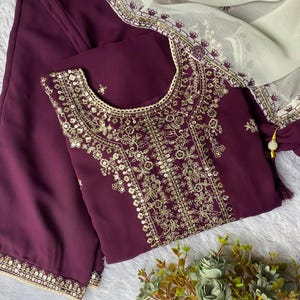 May include: A plum-colored embroidered tunic and matching pants are displayed with a cream-colored scarf. The tunic features intricate gold embroidery around the neckline and bodice. The scarf has a delicate gold border. A small bouquet of flowers is in the bottom right corner.