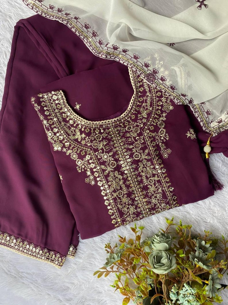 May include: A plum-colored embroidered tunic and matching pants are displayed with a cream-colored scarf. The tunic features intricate gold embroidery around the neckline and bodice. The scarf has a delicate gold border. A small bouquet of flowers is in the bottom right corner.