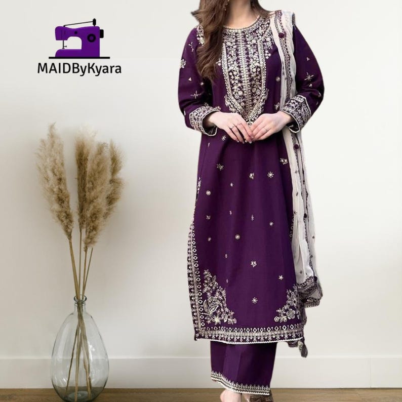 May include: A deep purple embroidered salwar kameez suit with matching pants and a cream-colored dupatta. The suit features intricate gold and silver embroidery around the neckline, sleeves, and hem. The brand logo "MAIDByKyara" is in the top left corner.