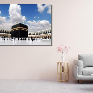 May include: A framed print of the Kaaba in Mecca, Saudi Arabia, under a bright blue sky with fluffy white clouds. The black cube-shaped structure is surrounded by a white marble courtyard and a colonnade.