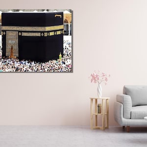 May include: A framed print of the Kaaba in Mecca, a black cube-shaped structure with gold accents, surrounded by a large crowd of people. The image is displayed on a wall in a room with a light pink wall, a gray sofa, and a small wooden table.