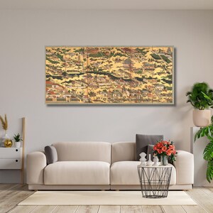 Könnte beinhalten: Ein großes, gerahmtes Kunstwerk, das eine detaillierte, historische Stadtlandschaft mit goldenen Akzenten zeigt. Das Kunstwerk ist über einem beigen Sofa angebracht, mit Pflanzen und einem Couchtisch davor. Das Kunstwerk zeigt Gebäude, Bäume und einen Fluss.