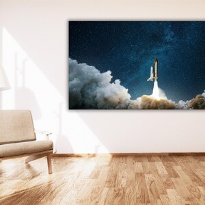 May include: A large canvas print depicting a space shuttle launching into a starry night sky. The shuttle is surrounded by billowing white smoke and flames. The artwork is mounted on a white wall above a wooden floor.