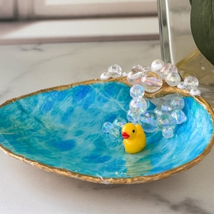 May include: A decorative, oval-shaped trinket dish with a blue and white marbled interior and gold-painted rim. A small yellow rubber duck figurine sits inside the dish, surrounded by clear bubble-shaped beads. Yellow flowers are in the background.