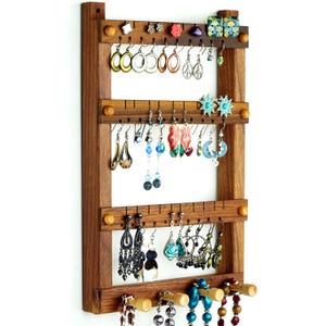 Könnte beinhalten: Holz-Ohrringhalter mit drei Ebenen mit Haken zum Aufhängen von Ohrringen. Der Halter hat eine braune Oberfläche und ist an einer weißen Wand montiert. An den Haken hängen mehrere Paar Ohrringe, darunter einige mit bunten Steinen und Perlen.