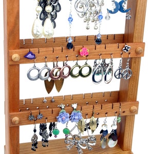 May include: A wooden earring stand with three tiers, each with multiple hooks for hanging earrings. The stand is made of light brown wood and has a natural finish. The stand is filled with a variety of earrings, including silver, gold, and colorful earrings.