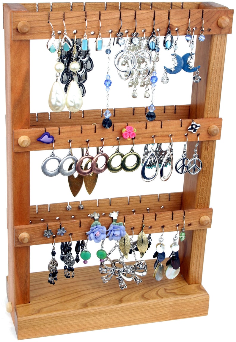 May include: A wooden earring stand with three tiers, each with multiple hooks for hanging earrings. The stand is made of light brown wood and has a natural finish. The stand is filled with a variety of earrings, including silver, gold, and colorful earrings.