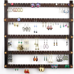 May include: A dark brown wooden earring holder with five rows of pegs. The holder has a rustic look and is filled with a variety of earrings, including silver, gold, and colorful stones.