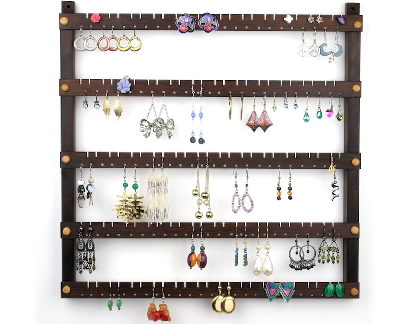 May include: A dark brown wooden earring holder with five rows of pegs. The holder has a rustic look and is filled with a variety of earrings, including silver, gold, and colorful stones.