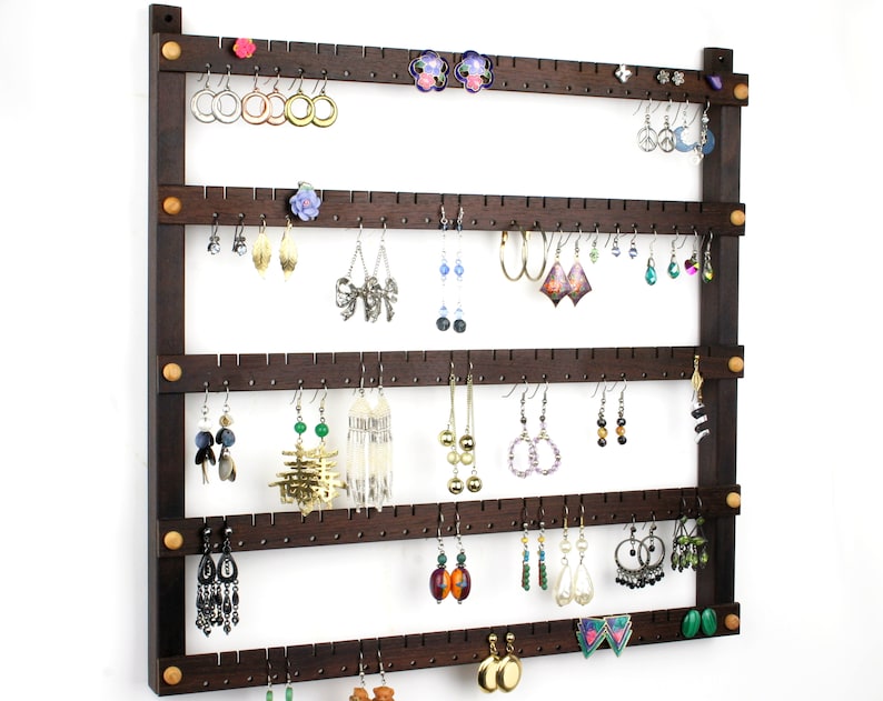 May include: A dark brown wooden earring holder with four rows of hooks. The holder has a total of 48 hooks and is mounted on a white wall. The holder is filled with a variety of earrings, including gold, silver, and colorful stones.