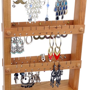 May include: A wooden earring holder with three tiers. The holder has multiple holes for hanging earrings. The holder is made of light brown wood and has a natural finish. There are a variety of earrings hanging on the holder, including silver, gold, and black earrings.
