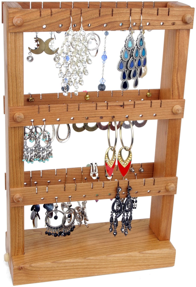 May include: A wooden earring holder with three tiers. The holder has multiple holes for hanging earrings. The holder is made of light brown wood and has a natural finish. There are a variety of earrings hanging on the holder, including silver, gold, and black earrings.