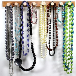 Puede incluir: Un organizador de joyas de madera con nueve collares colgados de ganchos. Los collares est&aacute;n hechos de varios materiales, incluyendo perlas, cuentas y cadenas met&aacute;licas. Los collares son de diferentes colores, incluyendo negro, blanco, verde, azul y marr&oacute;n.