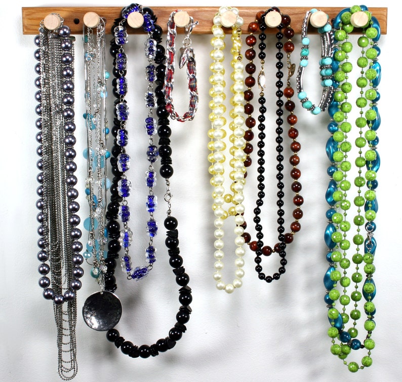 Puede incluir: Un organizador de joyas de madera con nueve collares colgados de ganchos. Los collares est&aacute;n hechos de varios materiales, incluyendo perlas, cuentas y cadenas met&aacute;licas. Los collares son de diferentes colores, incluyendo negro, blanco, verde, azul y marr&oacute;n.