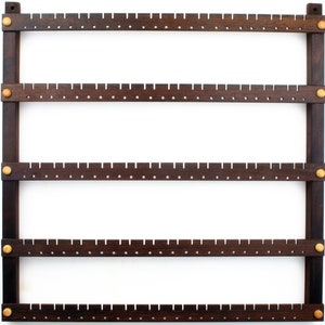 May include: A dark brown wooden earring holder with five rows of holes for storing earrings. The holder has gold metal accents at the corners and along the sides.