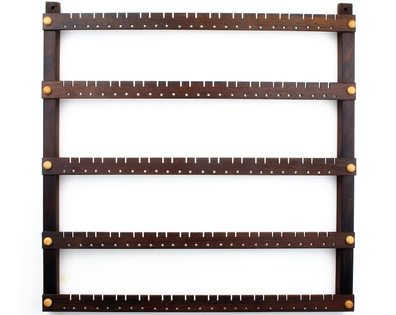 May include: A dark brown wooden earring holder with five rows of holes for storing earrings. The holder has gold metal accents at the corners and along the sides.