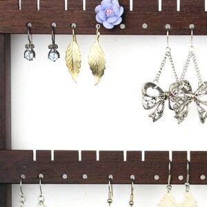 May include: A dark brown wooden earring holder with multiple rows of hooks. The holder has a small, light blue flower on the top row. There are several pairs of earrings hanging from the hooks, including silver bow earrings, gold leaf earrings, and silver earrings with clear stones.