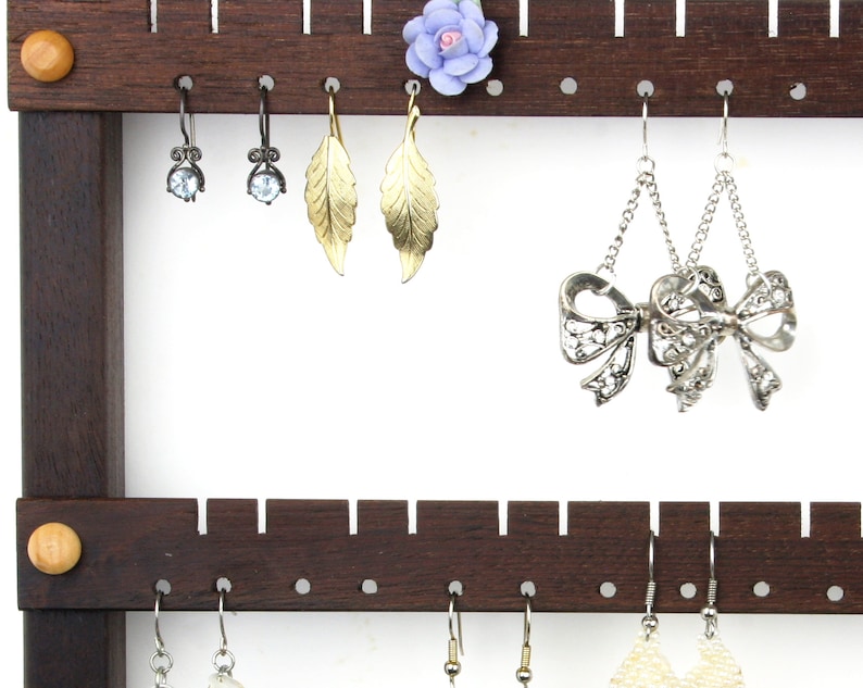 May include: A dark brown wooden earring holder with multiple rows of hooks. The holder has a small, light blue flower on the top row. There are several pairs of earrings hanging from the hooks, including silver bow earrings, gold leaf earrings, and silver earrings with clear stones.