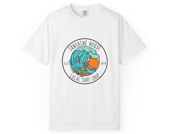 May include: White t-shirt featuring a circular graphic with a skeleton surfing a wave. The graphic includes the text "Sunshine House Local Surf Shop Est. 1979" in a black circular border. The design is in shades of blue, orange, and yellow.