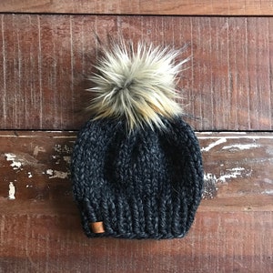 May include: A black knit beanie with a fluffy brown and white pom pom on top. The beanie has a brown leather tag on the side.