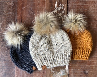 May include: Three knitted hats with faux fur pom poms. The hats are in black, white with black flecks, and mustard yellow.
