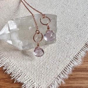 May include: A pair of delicate earrings with amethyst gemstone drops and gold-toned hoops. The earrings are displayed on a white crystal and a textured fabric surface.