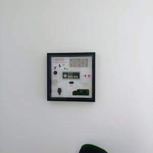 May include: A framed artwork featuring a deconstructed Nintendo controller design. The black frame surrounds a white background with various controller components and the Nintendo logo in red. The artwork is mounted on a white wall.
