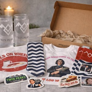 Puede incluir: Una colección de merchandising de Twin Peaks que incluye dos vasos transparentes con el logotipo de la serie, una camiseta blanca, calcetines, pegatinas y una caja de cartón. La camiseta tiene un gráfico de un personaje y el texto "LOG SOMETHING TO TELL".