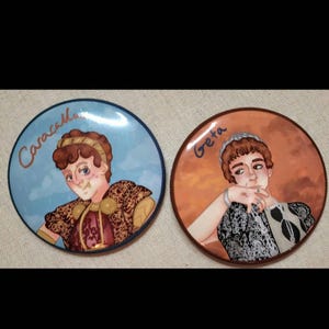 May include: Two circular ceramic plates with portraits. One plate shows a person named "Caracalla" in cursive. The other plate has a portrait of a person named "Geta". Both portraits are in a cartoon style, with a decorative border.