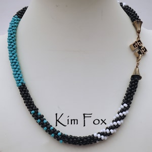 Puede incluir: Un collar de cuentas negras, blancas y turquesa con un cierre dorado. El collar está hecho de cuentas pequeñas y tiene un diseño geométrico. El collar está sobre un fondo blanco.