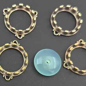 May include: Four bronze-colored metal ring components with gold-colored accents and a light blue, round stone. The rings have a decorative design with small, raised details around the outer edge. The stone has a hole in the center.
