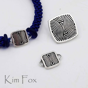 May include: A silver bracelet with a blue cord and silver square beads with a geometric pattern. The bracelet is shown with two additional silver square beads with the same geometric pattern.