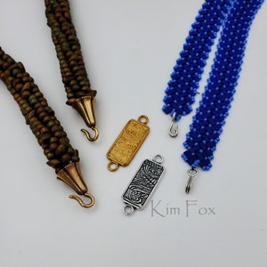 May include: Two beaded necklaces with different colored beads and patterns, each with a metal clasp. The necklaces are on a white surface with two metal rectangular clasps with decorative designs.