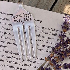 May include: A silver fork bookmark with the words "good book cozy nook" engraved on it. The fork is resting on a book with a sprig of lavender flowers.