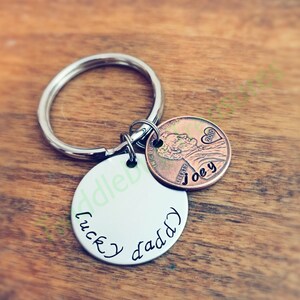 Pode incluir: Um chaveiro de prata com um charme redondo de prata que diz "lucky daddy" e uma moeda de cobre de um centavo com o ano 2010 e o nome "Joey" gravado nela.