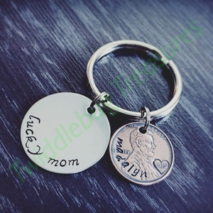 Pode incluir: Um chaveiro de prata com um charme redondo que diz "lucky mom" e um centavo de 2010 com as palavras "In God We Trust" e "Liberty" gravadas nele.
