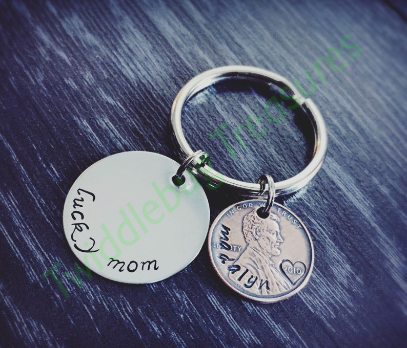Pode incluir: Um chaveiro de prata com um charme redondo que diz "lucky mom" e um centavo de 2010 com as palavras "In God We Trust" e "Liberty" gravadas nele.