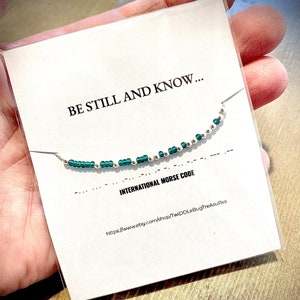 May include: A white card with the text "BE STILL AND KNOW..." and "INTERNATIONAL MORSE CODE" with a green and silver beaded bracelet on top of the card. The card also includes a website address.