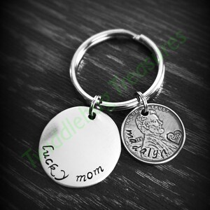 Pode incluir: Um chaveiro de prata com um charme redondo que diz "lucky mom" e um centavo de 2010 com as palavras "In God We Trust" e "Liberty" nele.
