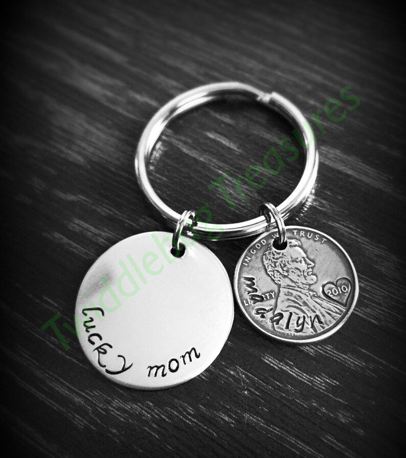Pode incluir: Um chaveiro de prata com um charme redondo que diz "lucky mom" e um centavo de 2010 com as palavras "In God We Trust" e "Liberty" nele.