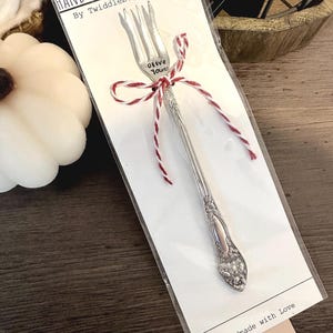 May include: A silver olive fork with the words "Olive You" stamped on the tines. The fork is tied with red and white twine and is packaged on a white card with the text "Hand-Stamped Olive Fork by Twiddlebug Treasures" and "Handmade with Love".