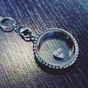 May include: A silver locket necklace with a clear glass insert and a silver heart charm with the initials "L&B" inside. The locket is surrounded by a row of clear rhinestones.