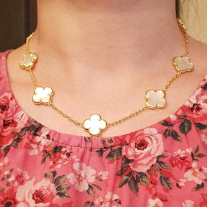 Puede incluir: Un collar dorado con colgantes blancos en forma de trébol. El collar está compuesto por una delicada cadena con seis abalorios de trébol. El fondo es un estampado floral rosa.