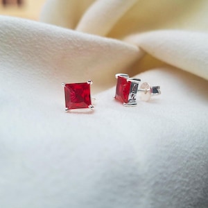 May include: A pair of square-cut, bright red gemstone stud earrings set in silver. The earrings are displayed on a soft, off-white fabric, showcasing their vibrant color and simple design. These earrings are a classic jewelry item.