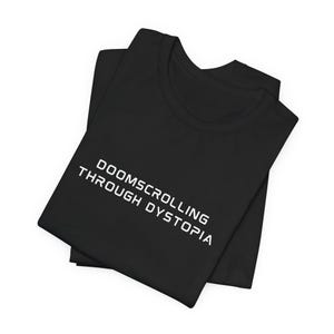 May include: Black t-shirt with the white text "DOOMSCROLLING THROUGH DYSTOPIA" printed across the front. The shirt is folded, showing the neckline and the printed design. The fabric appears to be soft and comfortable.
