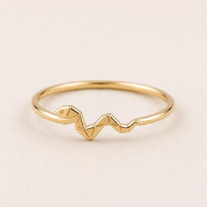 May include: A gold-toned ring featuring a thin, polished band with a geometric, faceted snake design integrated into the front. The snake motif has a segmented, angular appearance with a small circular indentation representing the eye.