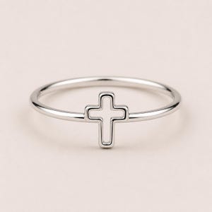 May include: A thin, polished silver-tone ring featuring a small, open-frame cross design centered on the band. The metal has a smooth, reflective finish, and the cross is integrated directly into the circular band, creating a continuous, minimalist look.