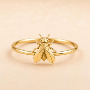 Può includere: Un anello color oro caratterizzato da un ornamento a forma di insetto geometrico centrale. L'insetto presenta un design angolare e sfaccettato con ali, corpo e antenne distinti. Il gambo dell'anello è sottile, liscio e circolare, creando un netto contrasto con il dettagliato elemento centrale sfaccettato.