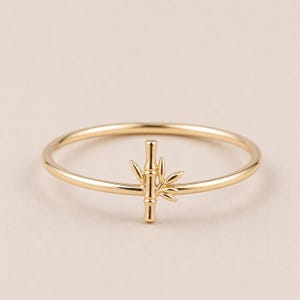 May include: A gold-colored ring featuring a bamboo design. The ring has a thin band and a detailed bamboo stalk with leaves as the focal point. The ring is set against a neutral background.